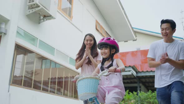 Asian couple father and mother teaching small pretty kid daughter in helmet riding on bike at street