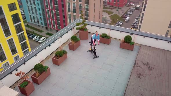 Healthy Caucasian Woman Exercising Workout on Stationary Cycling Machine Bike on House Rooftop Gym