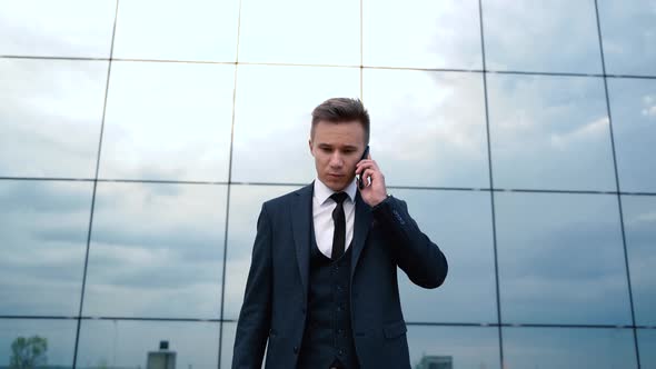 Handsome Young Caucasian Man Walking and Talking on Mobile Phone in City Center at Modern Building