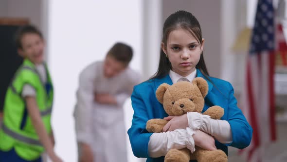 Sad Stressed Bullied Girl Hugging Teddy Bear As Boys Laughing Pointing at Background