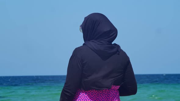 a Woman in a Hijab is Standing with Her Back to the Camera Against the Background of the Sea and