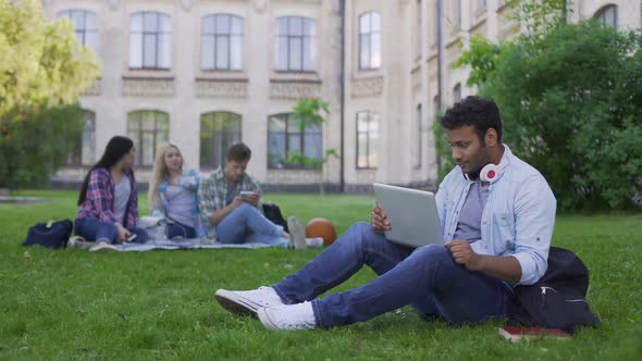 Mixed Race Student Chatting Online on Laptop, Sitting on Grass Near University