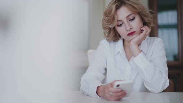 Portrait of Thoughtful Lady in White Shirt Uses Phone at Table in Light Home