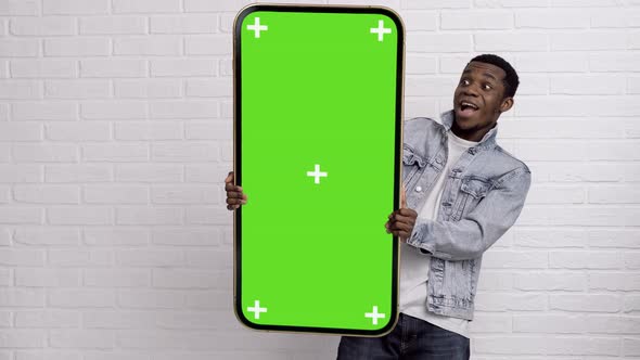 Excited Black Man Standing and Pointing Big Smartphone with Blank Green Screen Demonstrating Copy