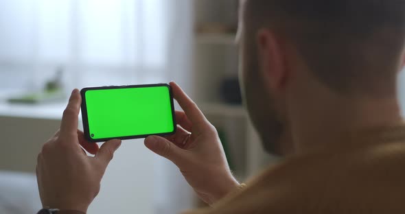 Man Is Viewing on Green Screen of Mobile Phone Holding Horizontally Modern Gadget and App for