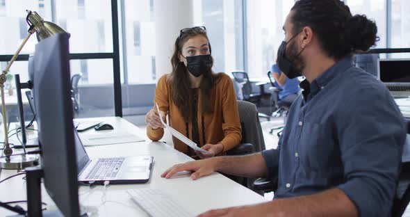 Diverse male and female colleague wearing face masks sitting at desk talking and elbow bumping
