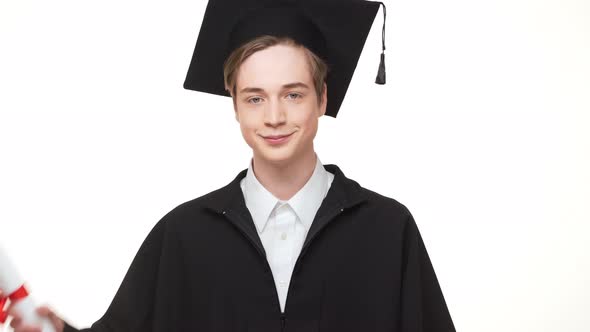 Happy Young Caucasian Male Graduate in Black Robe and Square Academic Cap Holding Scroll and Showing