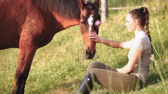 Young female sitting rider sitting on grass, stroking grazing horse