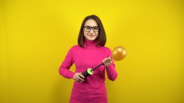 A Young Woman Inflates a Gold Balloon with a Pump on a Yellow Background. Girl in a Pink Turtleneck