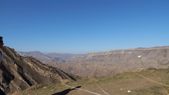 Young Man with a Backpack Walks Down the Slope Against the Background of Mountains on a Clear Autumn