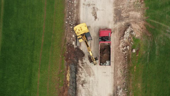 Top View Yellow Excavator Picks Up Land From the Field and Loads It Onto a Red Truck