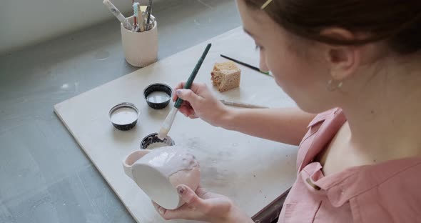 Female Potter Sitting and Stirs Paint with a Brush a Cup on the Table. Woman Making Ceramic Item