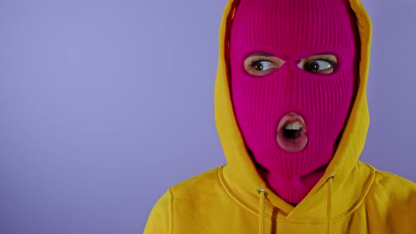 Close Up of Young Woman in Pink Balaclava and Yellow Hood Grimacing Face on Purple Background