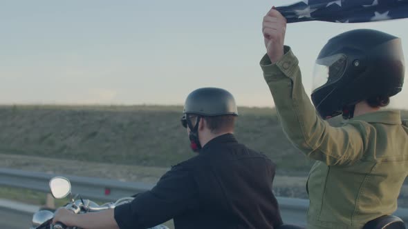 Couple in helmets rides motorbike on highway, woman holds an American flag flying over her head