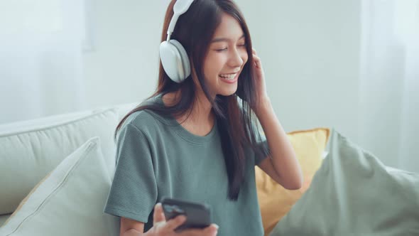 Happy young woman wear wireless headphones having fun listen music in smartphone and sit on sofa