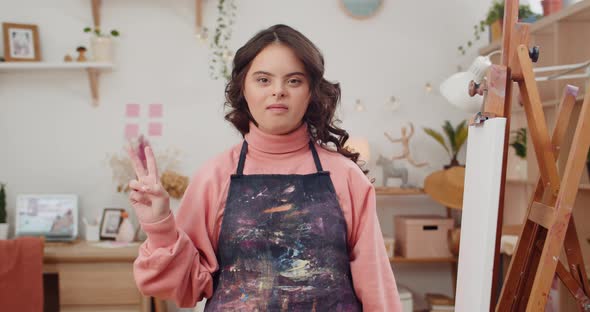 Adorable Teen with Down Syndrome Wearing in Artists Apron Showing Peace Sign While Looking To Camera