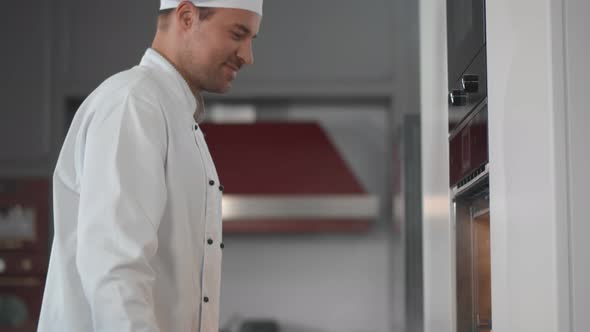 Young Man Baker Taking Fresh Croissants Out of Oven in Bakery Kitchen