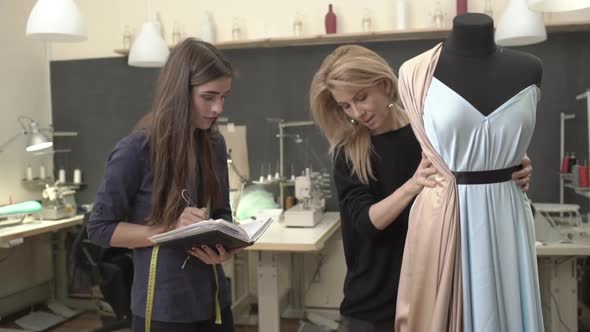 Blonde Caucasian Female Working with Mannequin with Azure Dress on It While Young Woman in Dark Blue