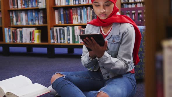 An Asian female student wearing a red hijab studying in a library and using tablet