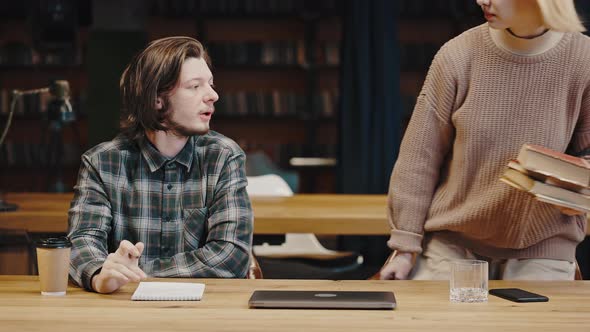 Young Pensive Man Studying with Notebook at Library Woman Groupmate Coming with Books and Giving One