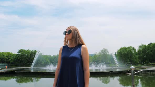 Steadicam Shot of a Beautiful Young Girl in Sunglasses in the Park Near the Fountain