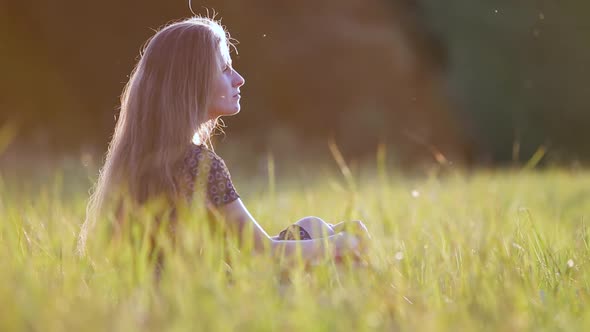 Young woman with long hair sitting outdoors in summer field grass enjoying nature at sunset.