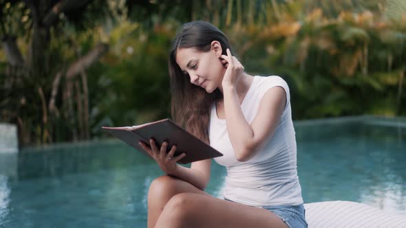 Young Woman in Summer Clothes Sits on Sunbed and Reads Book Near Pool