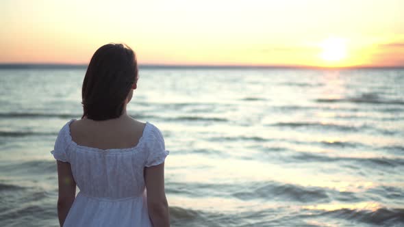 Attractive Young Woman Looks at the Sunset on the Beach By the Sea. The Girl in a White Dress Stands