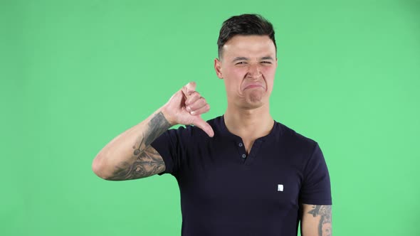 Portrait of a Handsome Young Guy Is Looking at Camera and Showing Thumbs Down Gesture. Green Screen