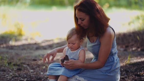 mother and baby sit in a park or in a forest on the ground looking at twigs and cones