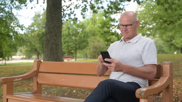 Happy Senior Man Sitting And Using Phone At The Park