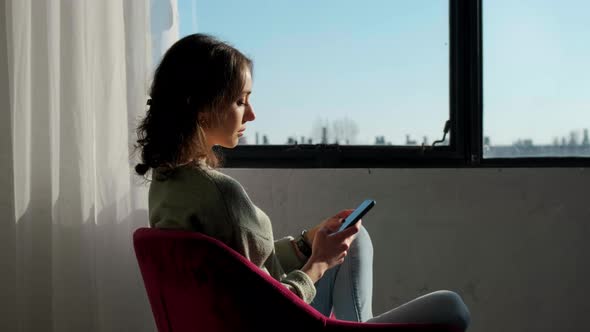 Beautiful Woman Sitting Near Window and Using Mobile Phone at Sunset