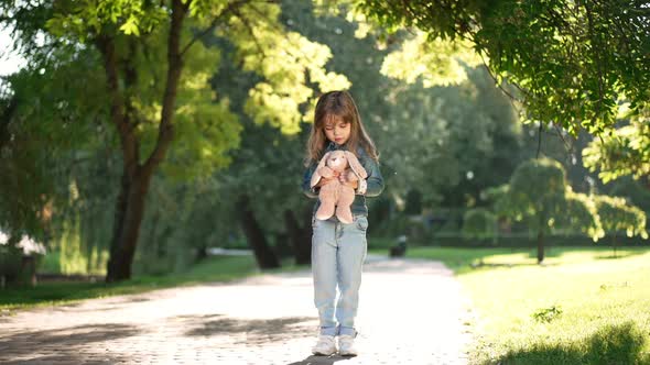 Wide Shot Portrait of Cute Caucasian Girl with Toy Standing in Sunshine in Spring Summer Park