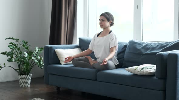 Young Indian woman sit on couch in lotus position in living room practice yoga