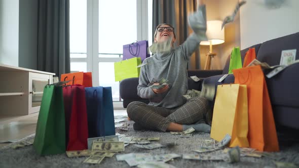 Happy Woman Is Sitting on a Carpet in a Cozy Room Among Shopping Bags and Making Money Rain From US