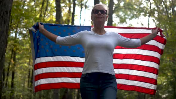 Backlit, pretty blonde woman with sunglasses walking through a forest holding a flag up in the air b