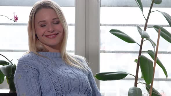 A Young Beautiful Woman Sits on a Chair By a Window and Smiles at the Camera