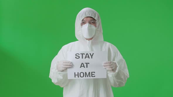 Asian Man Wear Protective Uniform PPE And Holding Stay At Home Sign In The Green Screen Studio