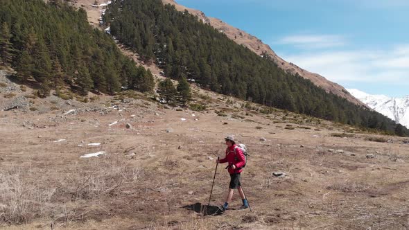 a Young Man in Shorts Sunglasses and a Cap with a Backpack Goes Along the Route in the Mountains