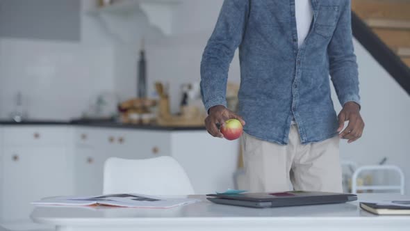 Young Professional Man Walking From Kitchen with Apple to Table in Home Office Sitting Down and