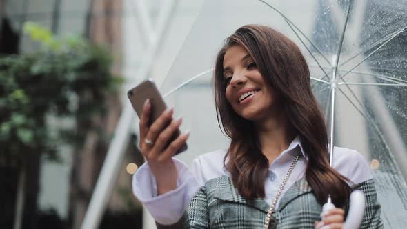 Beautiful Young Business Woman Using Smartphone on the Street in Rainy Weather, Smiling