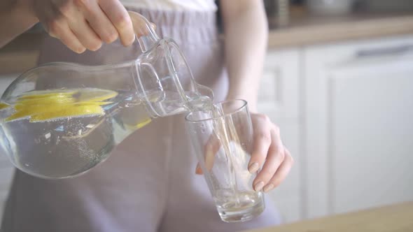 A Young Blonde Woman Pours Refreshing Water with Lemon Into a Transparent Glass and Drinks It