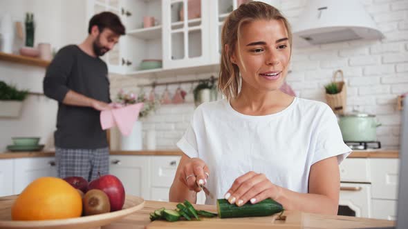 Young Woman Using Laptop and Preparing Vegatables for Salad While Young Bearded Man Wiping the