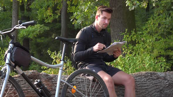 A Young Handsome Cyclist Sits on a Log Next To His Bike in a Forest and Reads a Map