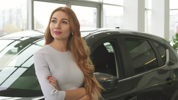 Joyful Young Woman Smiling Dreamily Leaning on a New Car at the Dealership