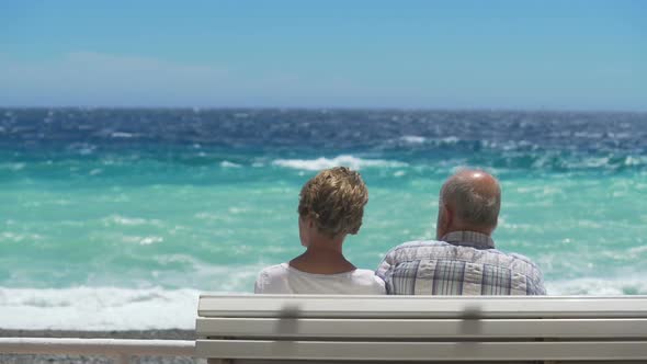 Aged Couple Sitting Next to Each Other on Bench by Waterfront, Facing Choppy Sea