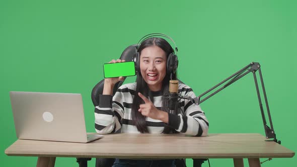 Asian Woman With Headphone Showing Green Screen Mobile Phone While Live Stream On Green Screen