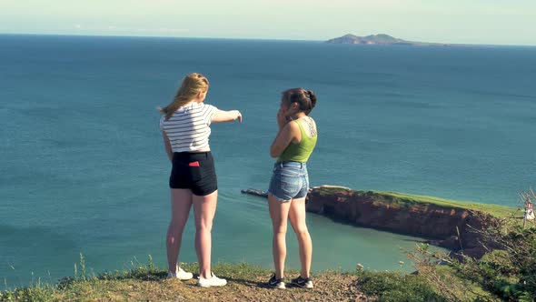 Two girls talk and gesture as they have a conversation near the edge of a tall cliff at the beach.