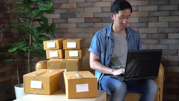 Asian Male Business Entrepreneur Using Laptop with Packs of Boxes at Home