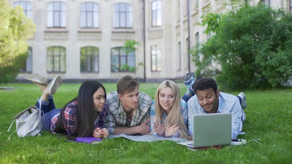 Group of Multi-Ethnic Students Relaxing on Grass and Watching Video on Laptop